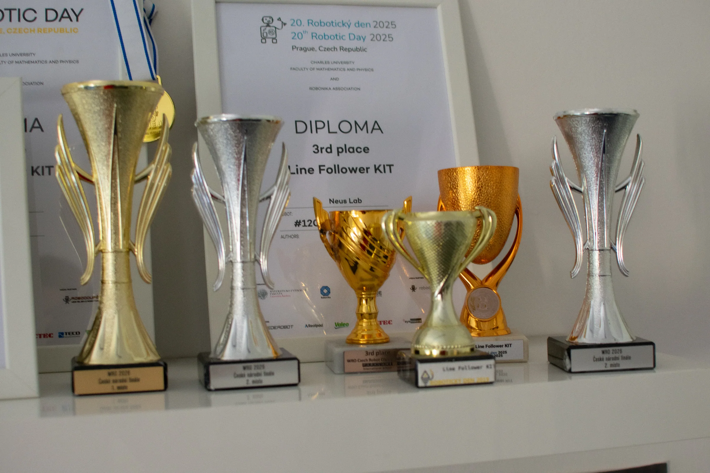 Skupina soutěžních pohárů NeusLab před zarámovanými diplomy